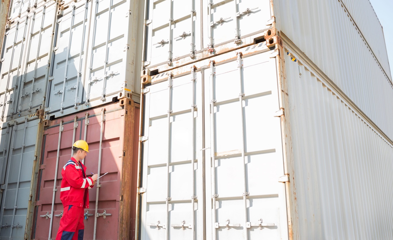 Worker inspecting containers while writing on clipboard in shipping yard